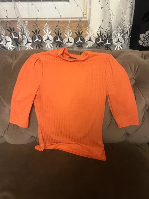 Ribbad orange topp med trekvartsärm - Snygg orange topp med ribbad struktur och trekvartsärm. Toppen har en högre hals och puffiga axlar som ger en trendig look. Perfekt att styla med jeans eller kjol för en färgstark outfit.