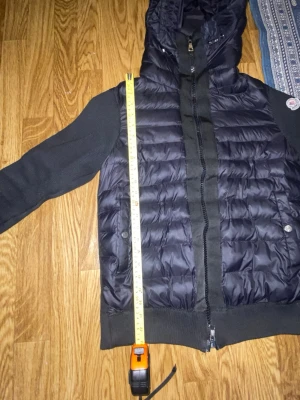 Monckler cardigan  - Snygg svart dunjacka från Moncler med quiltad front och stickade ärmar. Jackan har huva, dragkedja framtill och Moncler-logga på ärmen. Perfekt för kyliga dagar och ger en sportig men stilren look.