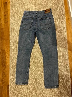 Blå Lee jeans med rak passform - Klassiska blå jeans från Lee med rak passform och fem fickor. Jeansen har en snygg mellanblå tvätt, Lee-lapp i läder bak i midjan och robusta sömmar. Perfekta för en avslappnad och stilren look.