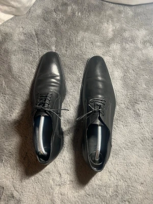 Svarta loafers från Hugo Boss i läder  - Stilrena svarta loafers från Hugo Boss i äkta skinn. Skorna har klassisk design med rund tå, diskreta sömmar och snörning framtill. Perfekta för dig som vill ha en clean och tidlös look passar för festligheter 