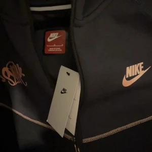 Central cee X Nike tech fleece zip up - Snygg svart hoodie från Nike med dragkedja framtill och orange Nike-logga på bröstet. Hoodien har huva, fickor med dragkedja och kontrastsömmar i orange. Perfekt för en sportig och avslappnad stil.