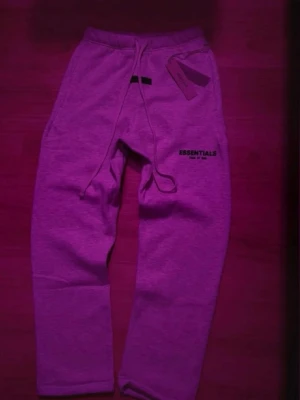 Grå Essentials mjukisbyxor med snörning - Säljer ett par grå mjukisbyxor från Essentials med snörning i midjan och tryckt logga på ena benet. Byxorna har en relaxed passform och är tillverkade i mjukt bomullsmaterial. Perfekta för chill dagar eller när du vill vara extra bekväm.