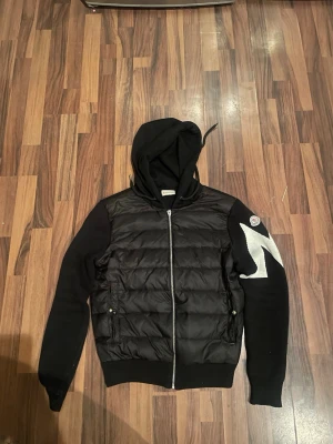 Svart teddyjacka med huva Moncler - Svart teddyjacka från Moncler med quiltad front, stickade ärmar och stor vit logga på vänster ärm. Jackan har huva med snörning och dragkedja framtill. Perfekt för dig som vill ha en stilren och sportig look.