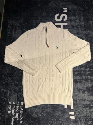 Vit kabelstickad tröja Polo Ralph Lauren - Kabelstickad vit tröja från Polo Ralph Lauren med halv dragkedja och ribbade muddar. Klassisk blå logga broderad på bröstet. Tröjan har hög krage och är tillverkad i mjukt material, perfekt för kyliga dagar.