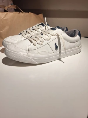 Vita sneakers från Polo Ralph Lauren - Snygga vita sneakers från Polo Ralph Lauren med klassisk logga i marinblått på sidan och mörkblå detaljer vid hälen. Skorna har rund tå, platt sula och snörning. Tillverkade i skinn med textilfoder för extra komfort.