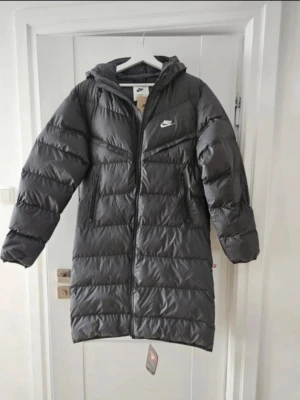 Svart lång dunjacka från nike  - Snygg svart dunjacka från Nike med huva och dragkedja framtill. Jackan är quiltad och har två praktiska sidofickor med dragkedja. Perfekt för kalla dagar och har en längre modell som håller dig varm hela vintern. Och storlek S