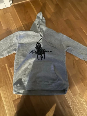 Grå hoodie från Ralph Lauren - Snygg grå hoodie från Ralph Lauren med stor svart polospelare och logga på bröstet. Klassisk känguruficka framtill och justerbar huva med dragsko. Perfekt för en avslappnad och trendig look. Mjuk bomullsblandning som känns skön mot huden. Pris kan diskuteras!
