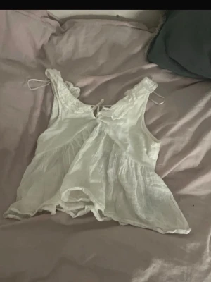 Vit volang topp med knyt - Söt vit topp med volangdetaljer och knyt i ryggen. Gjord i ett tunt, luftigt tyg som känns perfekt för varma dagar. Ärmlös modell med lätt rynkad struktur och romantisk känsla. Passar dig som gillar en feminin och avslappnad stil.