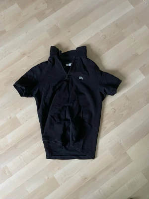Svart pikétröja från Lacoste - Klassisk svart pikétröja från Lacoste med korta ärmar och den ikoniska krokodilloggan på bröstet. Tröjan har krage och knäppning framtill. Tillverkad i mjukt bomullsmaterial som känns skönt mot huden. Säljer pga att den är för liten! Pris inte hugget i sten