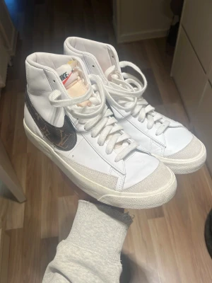 Nike Blazer Mid med brun custom detalj - Snygga vita Nike Blazer Mid sneakers med högt skaft och custom-made detaljer. Skorna har klassisk snörning, rund tå och är gjorda i skinn och syntet. Perfekta för dig som vill sticka ut med en unik look.