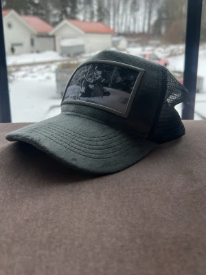 Grå truckerkeps med hockeymotiv - Säljer en grå truckerkeps med svart mesh baktill och ett stort patch framtill med hockeymotiv och texten 'NBCHICA TIARIS'. Kepsen har böjd skärm sopassad för en avslappnad streetstil.