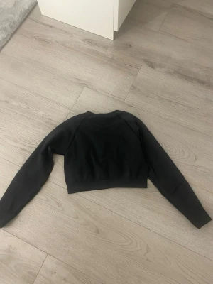 Svart croppad träningtröja från xshape - Svart croppad träningtröja från xshape med rund halsringning och långa ärmar. Tröjan har en enkel och stilren design utan mönster eller detaljer, perfekt för en avslappnad look. Tillverkad i mjuk bomullsmix för extra komfort.