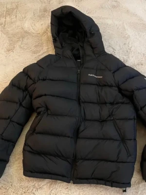 Svart dunjacka från Peak Performance - Snygg svart dunjacka från Peak Performance med huva och dragkedja framtill. Jackan har en quiltad design och är perfekt för kalla dagar. Klassisk pufferstil med logga på bröstet och praktiska fickor på sidorna.