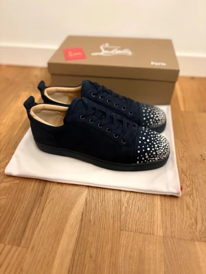 Marinblå sneakers med nitar från Louboutin - Snygga marinblå sneakers från Christian Louboutin i mocka med ikonisk röd sula och silverfärgade nitar på tån. Skorna har snörning och rund tå, samt en lyxig känsla med detaljer som sticker ut.
