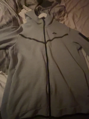 Grå Nike hoodie med dragkedja - Säljer en grå hoodie från Nike med hel dragkedja framtill och svart Nike-logga på bröstet. Hoodien har huva och svarta detaljer längs dragkedjan och axlarna. Perfekt för en sportig och avslappnad stil.