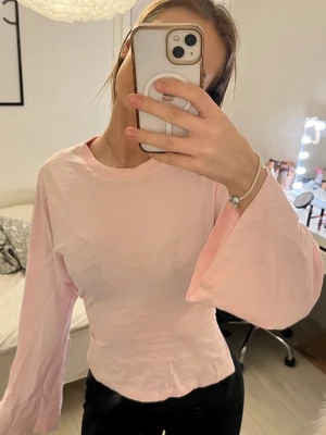 Ljusrosa omlottopp med knyt i ryggen - Supersöt ljusrosa topp med långa vida ärmar och snygg omlottdetalj i ryggen som knyts. Toppen är croppad och har en mjuk, tunn känsla. Perfekt för dig som gillar trendiga detaljer och en feminin look.