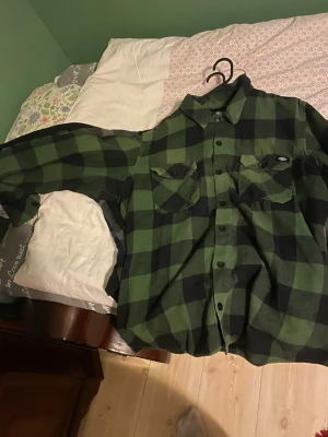 Grön och svart rutig skjorta Dickies - Säljer en grön och svart rutig skjorta från Dickies med klassisk krage, två bröstfickor med lock och knappar framtill. Skjortan har lång ärm och är tillverkad i ett mjukt material som passar perfekt för en avslappnad stil.