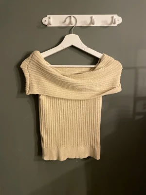 Ribbad offshoulder topp i beige - Trendig ribbad offshoulder topp i beige med bred krage som vilar över axlarna. Toppen är stickad och har en kroppsnära passform som framhäver siluetten. Perfekt för dig som vill ha en stilren och modern look. Tryck köp nu❤️