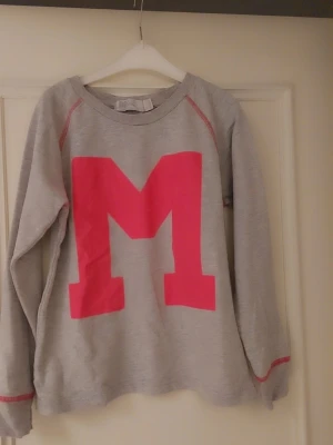 Grå sweatshirt med rosa M - Säljer en grå sweatshirt med en stor rosa bokstav M på bröstet. Tröjan har rund halsringning, långa ärmar och rosa sömdetaljer vid axlar och ärmslut. Perfekt för dig som gillar en sportig och avslappnad stil.