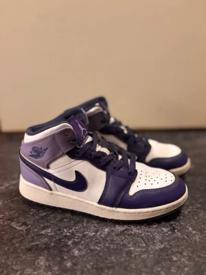 Nike Air Jordan 1 Mid  - Nike Air Jordan 1 Mid sneakers i lila och vitt med klassisk snörning och perforerad tå. Skorna har en ikonisk Swoosh på sidan och Air Jordan-logga vid ankeln. Tillverkade i skinn med kontrasterande paneler och rund tå. Perfekta för dig som gillar streetwear.