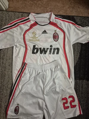 AC Milan Kaka matchställ Adidas Size M - Säljer ett vitt AC Milan matchställ från Adidas med Kaka och nummer 22 på ryggen. Tröjan har röda och svarta detaljer, Champions League-märke och Bwin som sponsor. Shortsen har samma färgtema och nummer 22. Perfekt för fotbollsfans!