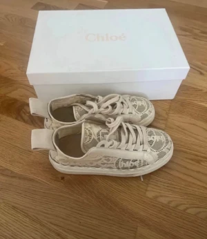 Beige spets-sneakers från Chloé - Säljer ett par snygga beige sneakers från Chloé med spetsdetaljer och tydlig logga på sidan och hälen. Skorna har snörning och platt sula, samt en stor tygflik bak med Chloé-tryck. Perfekta för dig som gillar unika och trendiga detaljer. Har ej kommit till användning 