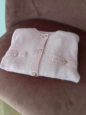 Ljusrosa kortärmad kofta med pärlknappar - Supersöt ljusrosa kofta med korta ärmar och rund hals. Koftan har dekorativa fickor framtill och pärlknappar i guld-look längs hela framsidan. Ribbstickade kanter vid ärmslut, hals och nederkant. Perfekt för en söt och trendig look. Använd en gång så är som helt ny, ina märken på användning syns.