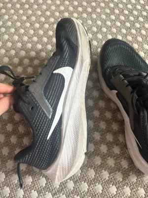 Svarta Nike sneakers med vit sula - Snygga svarta Nike sneakers med vit sula och stor vit swoosh på sidan. Ovandelen är i mesh-liknande syntetmaterial för bra andningsförmåga. Klassisk snörning och låg profil gör dem perfekta för en sportig look.