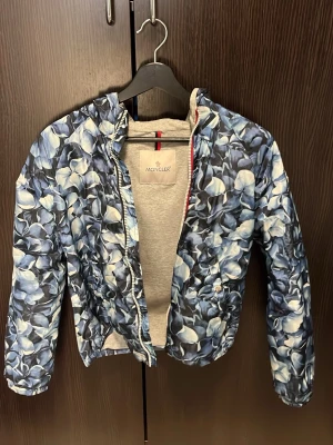 Blommig vindjacka från Moncler - Snygg vindjacka från Moncler med blått och vitt blommönster. Jackan har dragkedja framtill, huva och långa ärmar. Tillverkad i lätt polyester som passar perfekt för vår och sommar. Perfekt för dig som vill sticka ut med en unik jacka.