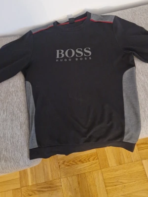 Svart sweatshirt från Hugo Boss - Snygg svart sweatshirt från Hugo Boss med grå paneler på sidorna och diskret röd detalj vid axlarna. Stor BOSS-logga framtill. Perfekt för en avslappnad och trendig look. Mjuk bomullsblandning och rund hals.