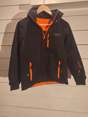 Svart vindjacka från Swedemount - Svart vindjacka från Swedemount med orange detaljer och dragkedjor. Jackan har huva, flera fickor med dragkedja och logga på bröstet. Insidan är fodrad i orange och jackan har lång ärm. Perfekt för blåsiga dagar.