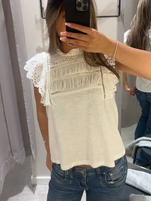 Vit blus med spets från Holly & Whyte - Säljer en söt vit blus från Holly & Whyte med broderade spetsdetaljer och volang vid axlarna. Blusen har ärmlös design och rundad hals med små volanger.köpt för 499kr och är använd fåtal gånger, skriv ifall du undrar något  