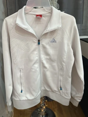 Vit ziptröja från Peak - Snygg vit ziptröja från Peak med diskret mönster på bröstet och blå detaljer på dragkedjorna. Tröjan har hög krage, två fickor med dragkedja och ribbade muddar. Perfekt för en sportig och avslappnad stil. Men är tyvär smutsigt, den har testat att bli tvättad men är fast.