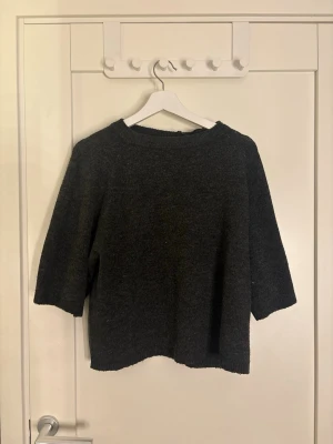 Mörkgrå stickad tröja med trekvartsärm - Mysig mörkgrå stickad tröja med trekvartsärmar och rund halsringning. Tröjan har en rak passform och är perfekt för lager-på-lager-stil. Materialet ser ut att vara ull eller ullblandning.