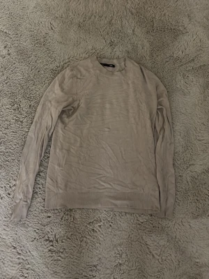 Beige stickad tröja med rund hals - En stilren beige stickad tröja med rund halsringning och långa ärmar. Tröjan har ribbstickade muddar vid ärmslut och nederkant. Perfekt för lager-på-lager och enkel att matcha med olika outfits.