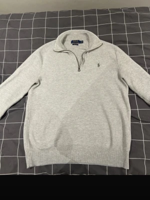 Ljusgrå stickad tröja Polo Ralph Lauren - Ljusgrå stickad tröja från Polo Ralph Lauren med halv dragkedja och hög krage. Tröjan har lång ärm, ribbstickade muddar och den klassiska broderade loggan på bröstet. Perfekt för lager-på-lager och en clean look. Storlek S och är i perfekt skick.