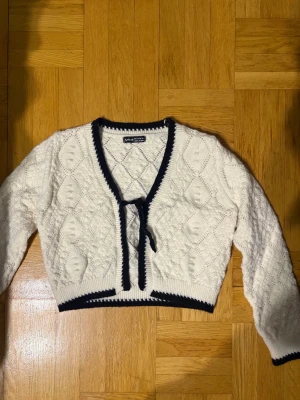 Vit cropped kofta med svarta detaljer - Säljer en vit cropped kofta från SHEIN med svart kantband och knytning framtill. Koftan har ett stickat mönster med diskreta romber och långa ärmar. Perfekt för att lyfta en outfit med en söt och trendig touch.