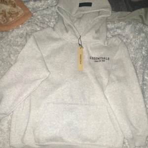 Grå Essentials Fear of God hoodie - Snygg grå hoodie från Essentials Fear of God med svart logga tryckt på bröstet. Klassisk modell med huva och känguruficka framtill. Mjuk bomullsblandning som är perfekt för en avslappnad streetwear-look.