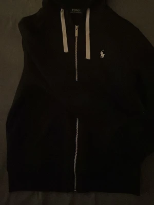 Svart zip hoodie från Polo Ralph Lauren - Snygg svart hoodie med dragkedja från Polo Ralph Lauren. Klassisk vit broderad logga på bröstet och vita snören i huvan. Perfekt för en avslappnad och stilren look. Tillverkad i mjukt material för extra komfort. Säljer eftersom den ej passar längre