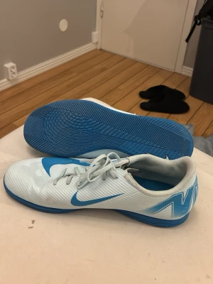 Nike vita och blå fotbollsskor - Säljer ett par vita och blå fotbollsskor från Nike med klassisk snörning och låg profil. Skorna har en blå yttersula och stora blå Nike-loggor på sidorna. Tillverkade i syntetmaterial för bra grepp och känsla på planen.