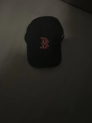 Svart Boston Red Sox keps - Säljer en svart keps från New Era med Boston Red Sox-loggan broderad i rött och vitt framtill. Klassisk böjd skärm och justerbar passform bak. Perfekt för dig som gillar sportig streetstyle och vill ha en ikonisk look.