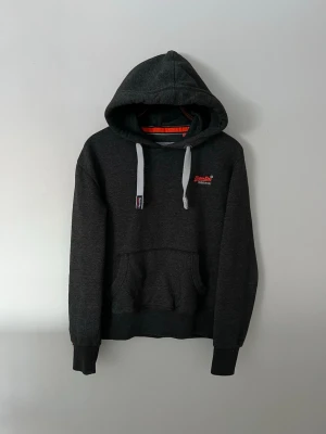 Superdry Hoodie - Snygg mörkgrå hoodie från Superdry med broderad logga i rött och vitt på bröstet. Hoodien har stora fickor framtill, gråvita dragsnören och en klassisk huva. Tillverkad i mjukt bomullsmaterial som känns skönt mot huden. Perfekt för en chill och avslappnad stil.