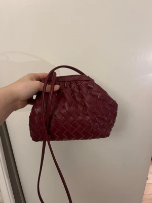 Vinröd flätad clutch väska  - Säljer en vinröd flätad clutchbag med lång axelrem. Väskan har ett rymligt fack med dragkedja på insidan.