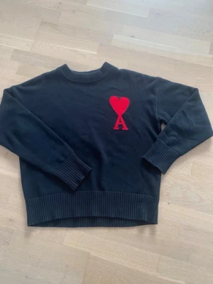 Svart stickad tröja från Ami Paris - Svart stickad tröja från Ami Paris med röd hjärtlogga och bokstaven A på bröstet. Tröjan har rund hals, ribbade muddar och nederkant. Perfekt för dig som gillar stilrena plagg med en twist.