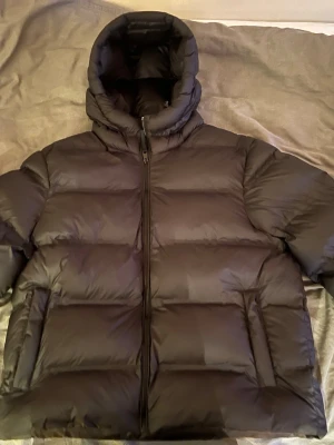 Svart pufferjacka med huva - Snygg svart pufferjacka med hög krage och stor huva. Jackan har quiltad design, dragkedja framtill och två sidofickor. Perfekt för kalla dagar och ger en riktigt modern look. Passformen är normal och materialet känns som syntet.