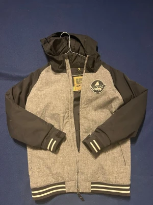 Grå och svart vindjacka från Burton - Snygg vindjacka från Burton i grått och svart med huva och dragkedja. Jackan har ribbade muddar med vita ränder och ett stort Burton-märke på bröstet. Perfekt för dig som vill ha en sportig och stilren look.