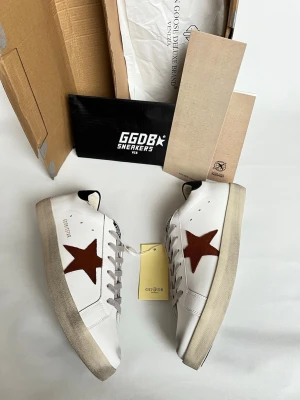 Golden Goose vita sneakers med stjärna - Golden Goose sneakers i vitt skinn med brun stjärndetalj på sidan och grå snörning. Klassisk låg modell med rund tå och platt sula. Märkeslogga på plösen och bak på hälen. Snygg och tidlös design som passar till det mesta.