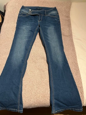 Blå bootcut jeans från Midross - Snygga blå lågmidjade bootcut jeans från Midross med bred linning och tre silverfärgade knappar. Jeansen har klassiska fickor fram och bak, samt markerade sömmar. Perfekta för dig som gillar en retroinspirerad look med utsvängda ben. Säljer för att dom inte passar på mig. Dom är helt oanvända.