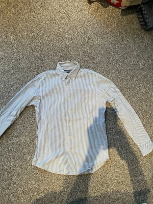 Randig skjorta från Ralph Lauren - Klassisk skjorta från Ralph Lauren med tunna blå och vita ränder. Skjortan har lång ärm, knappar framtill och en liten rosa logga broderad på bröstet. Perfekt till jeans eller chinos för en clean och stilren look.