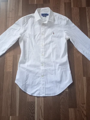 Vit skjorta från Ralph Lauren - Klassisk vit skjorta från Ralph Lauren i slim fit-modell. Skjortan har långa ärmar, knappar framtill och den ikoniska broderade loggan på bröstet. Tillverkad i bomull för en skön och stilren känsla.